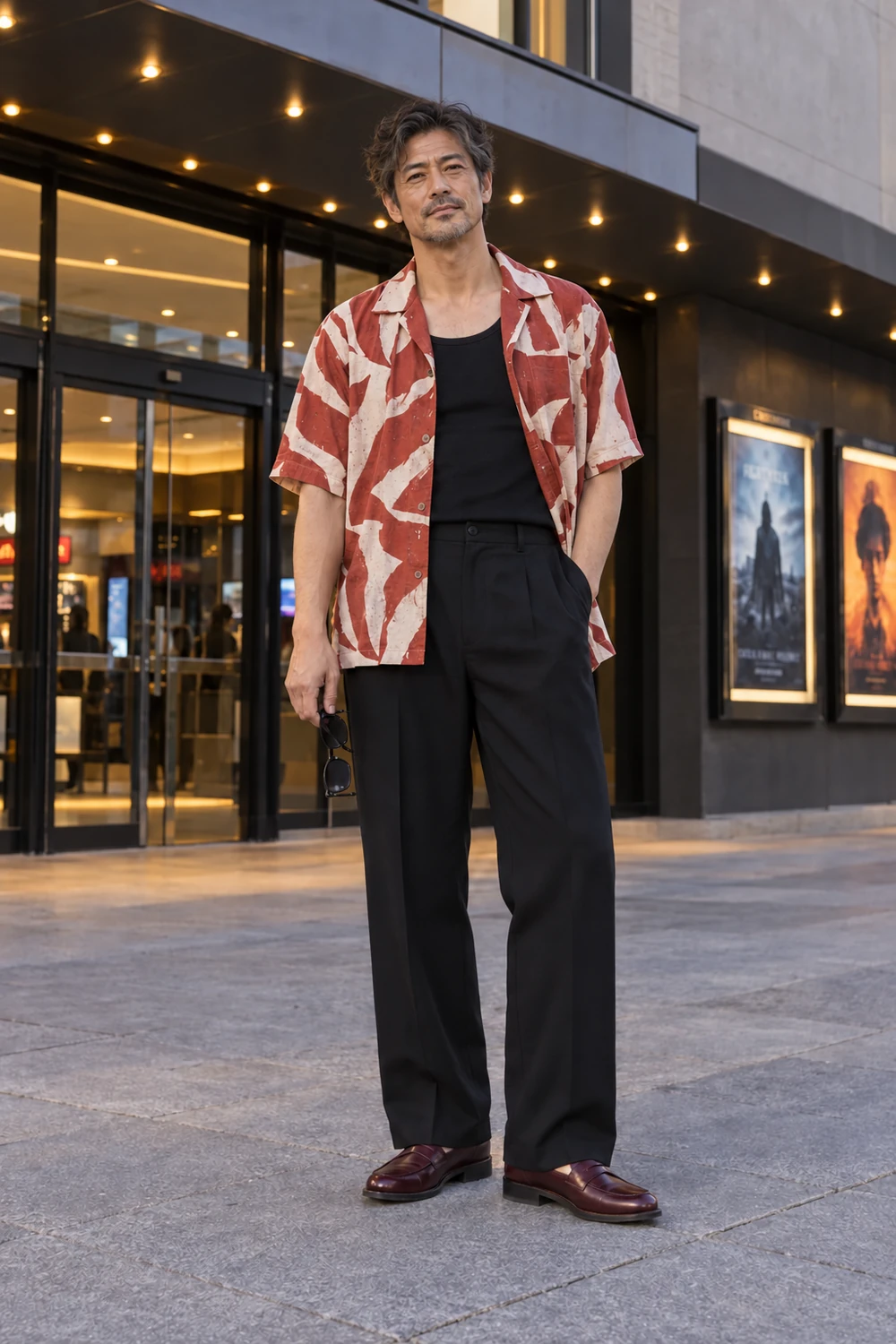A lean mature fictional man with light-medium warm skin and short salt-and-pepper hair wearing an open brick-and-cream printed shirt over a black top, black wide trousers and burgundy low loafers, holding dark glasses, shown full length.