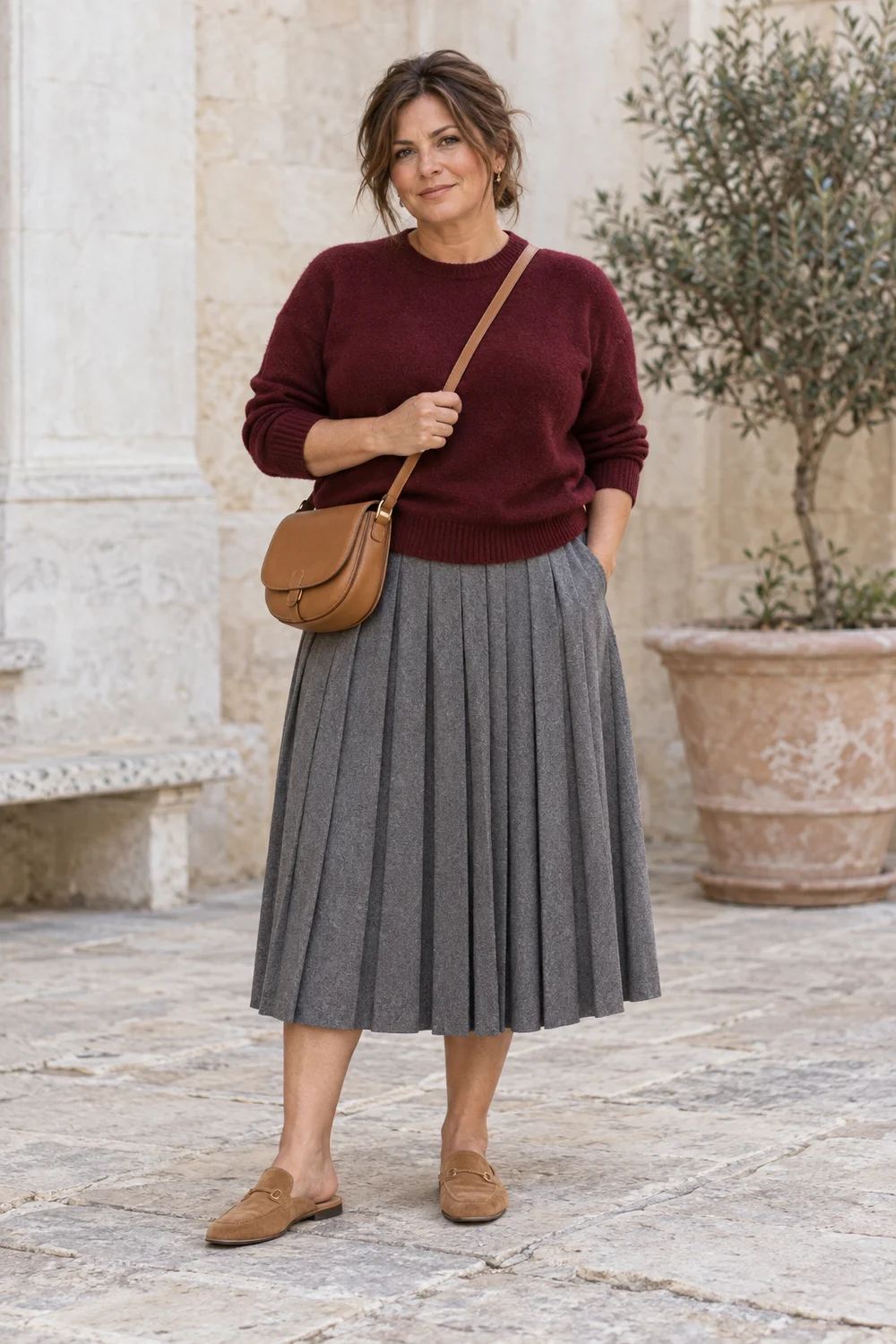A fictional woman in grey pleated midi skirt, burgundy knit, tan flat mules and tan crossbody bag.