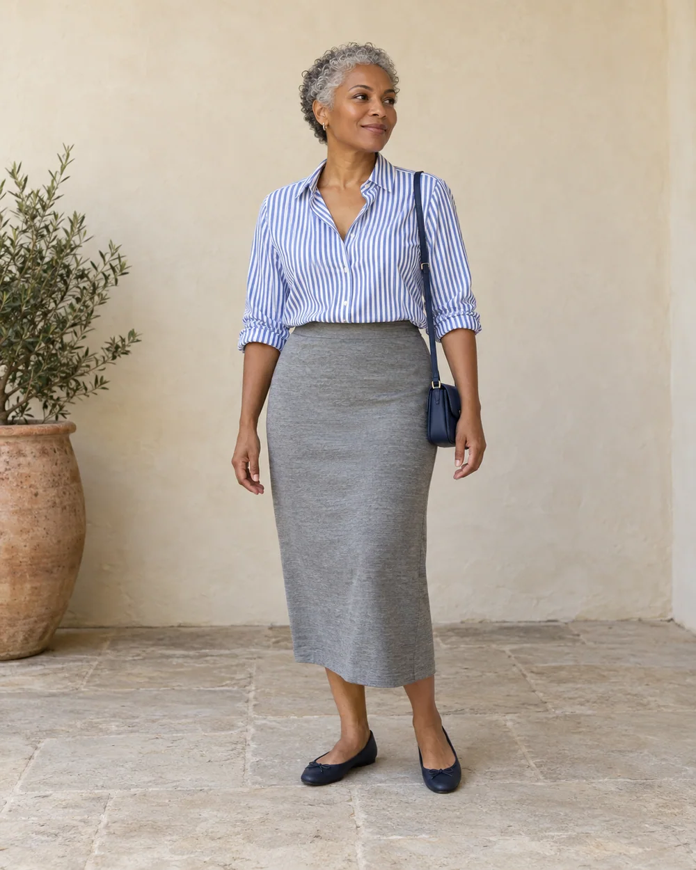 A full-length adult with medium-deep warm brown skin wearing Grey jersey midi skirt, blue-and-white striped shirt and Navy ballet flats, with a compact navy bag; both complete shoes are visible.