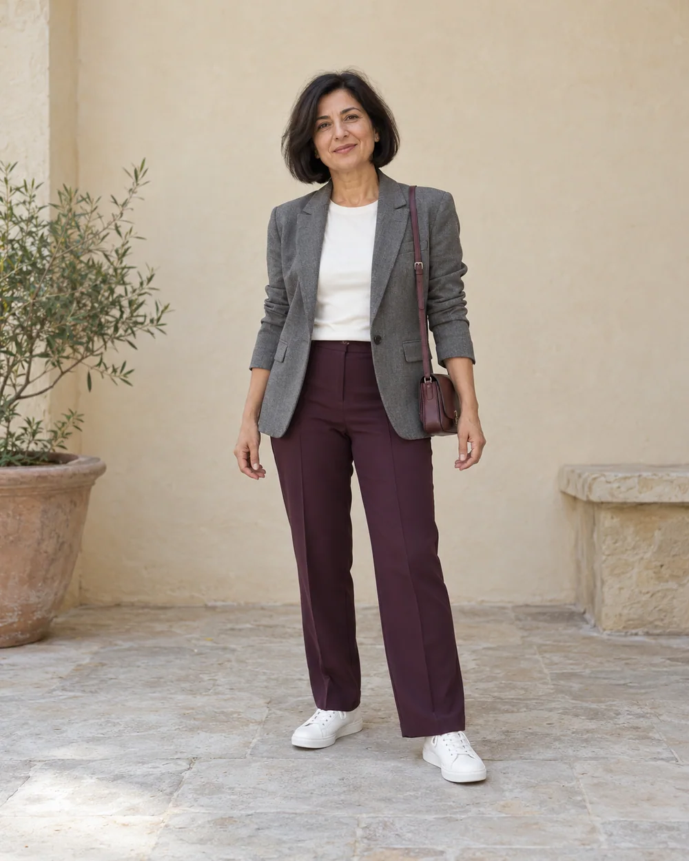 A full-length adult with medium olive skin wearing Plum straight trousers, ivory opaque tee, Grey relaxed blazer and White leather trainers, with a compact burgundy bag; both complete shoes are visible.