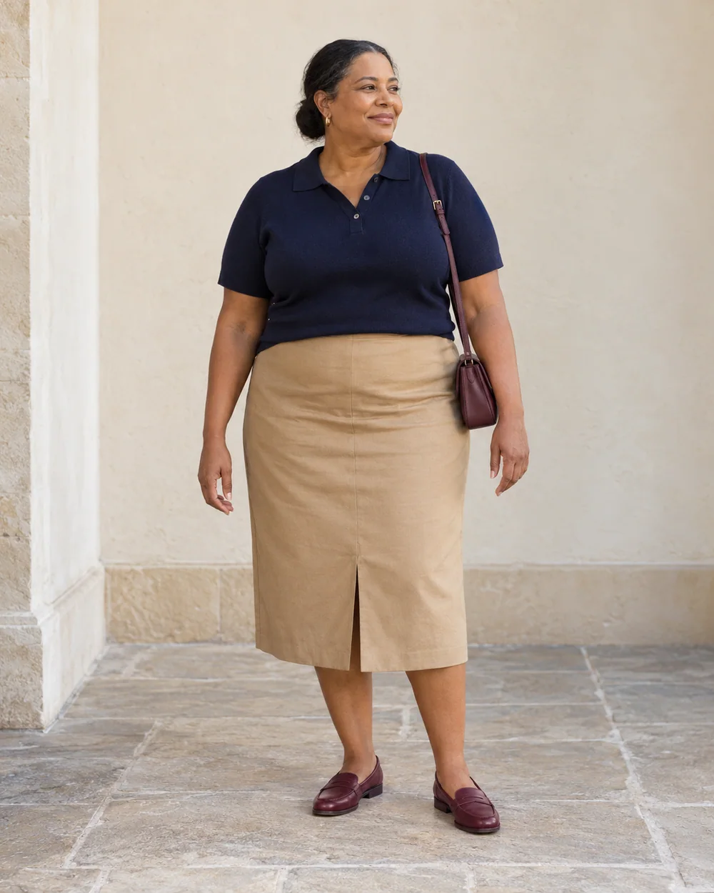 A full-length adult with medium-deep warm brown skin wearing Tan straight skirt with walking vent, navy knit polo and Burgundy flat loafers, with a compact burgundy bag; both complete shoes are visible.