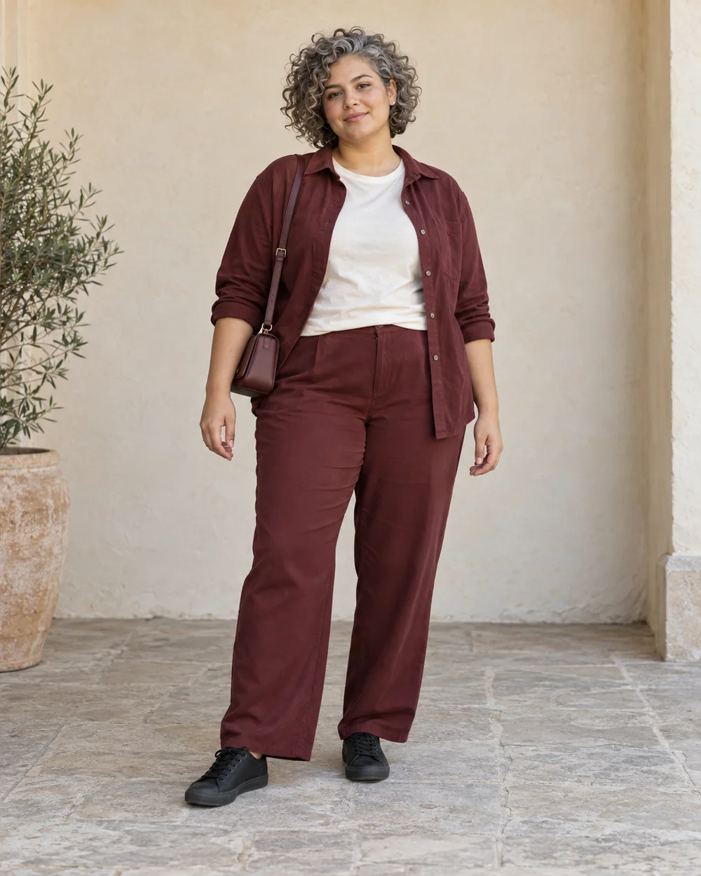 A full-length adult with medium olive skin wearing Burgundy relaxed trousers, ivory tee, Burgundy open overshirt and Plain black low trainers, with a compact burgundy bag; both complete shoes are visible.