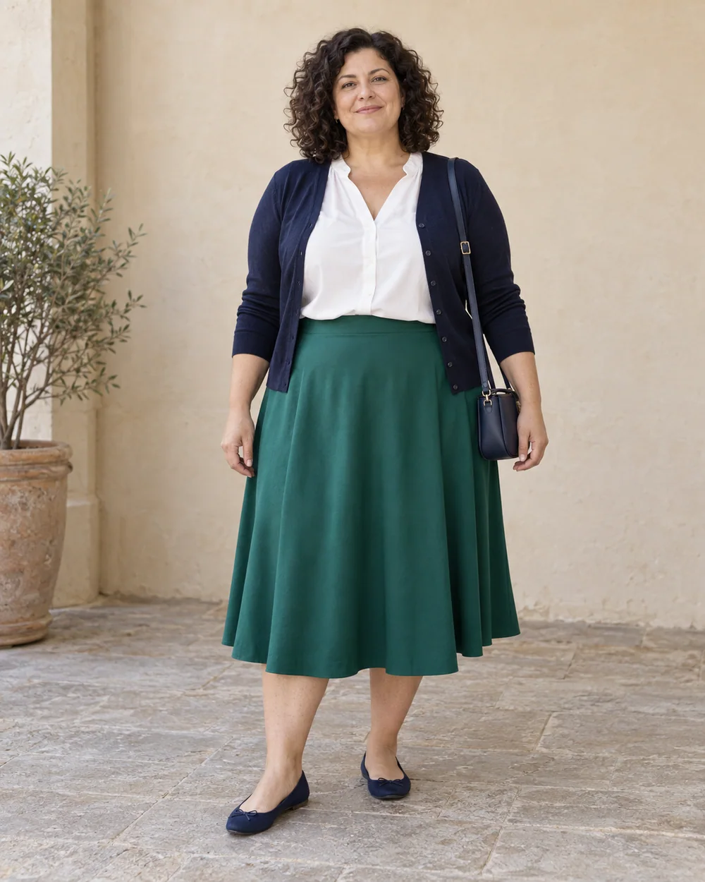 A full-length adult with light-medium neutral skin wearing Emerald A-line midi skirt, opaque white blouse, Navy fine cardigan and Navy ballet flats, with a compact navy bag; both complete shoes are visible.