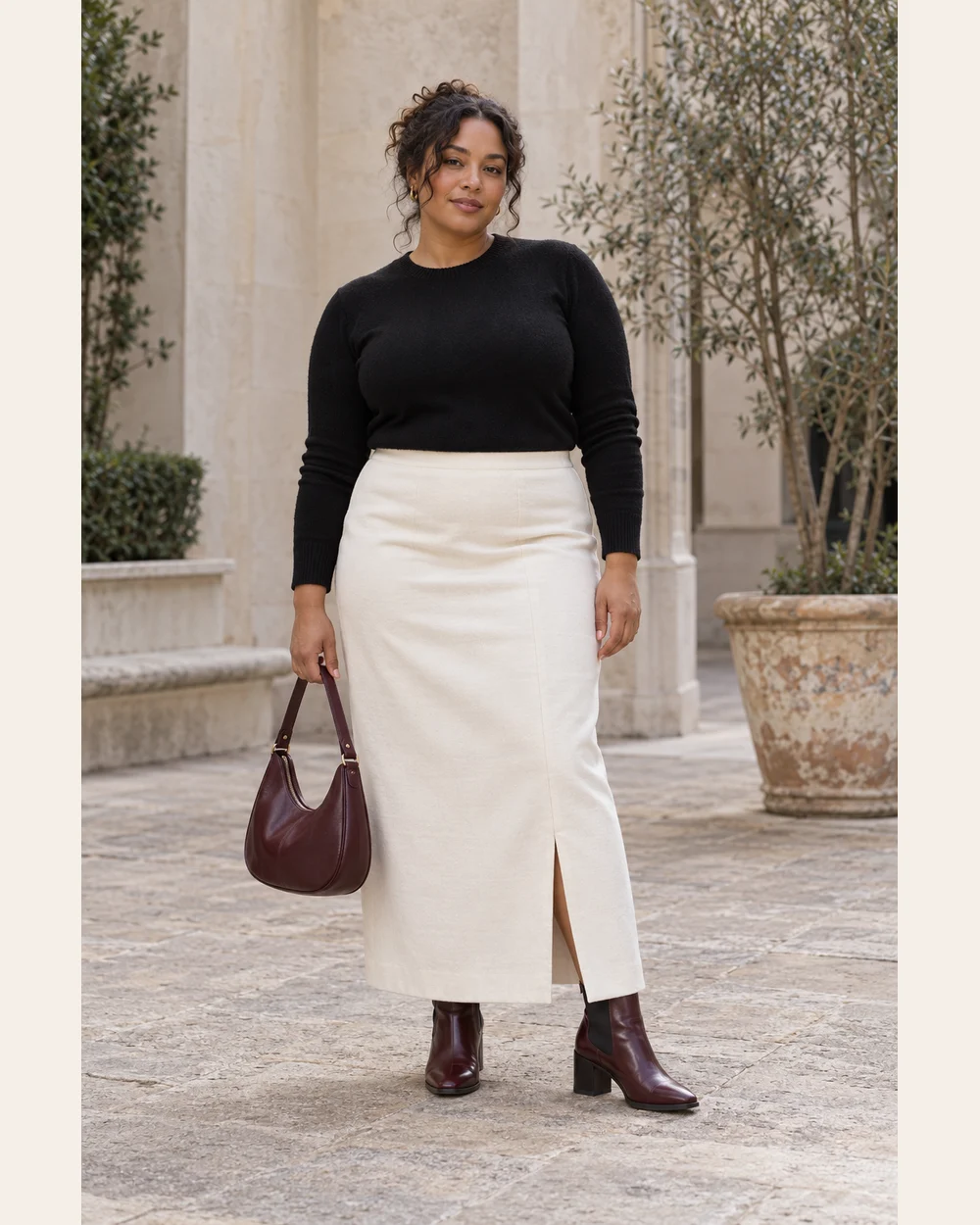 A fictional woman in ivory column skirt, black fine knit, oxblood heeled Chelsea boots and burgundy bag.