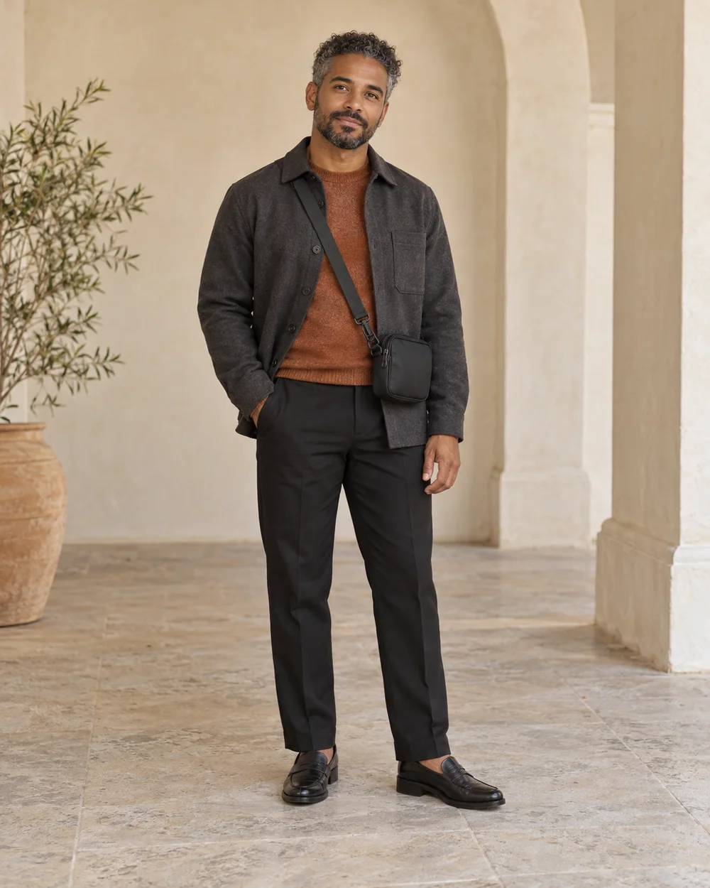 A full-length adult man with medium-deep warm brown skin wearing Black straight trousers, copper fine knit, Charcoal overshirt and Black loafers, with a small dark crossbody bag; both complete shoes are visible.