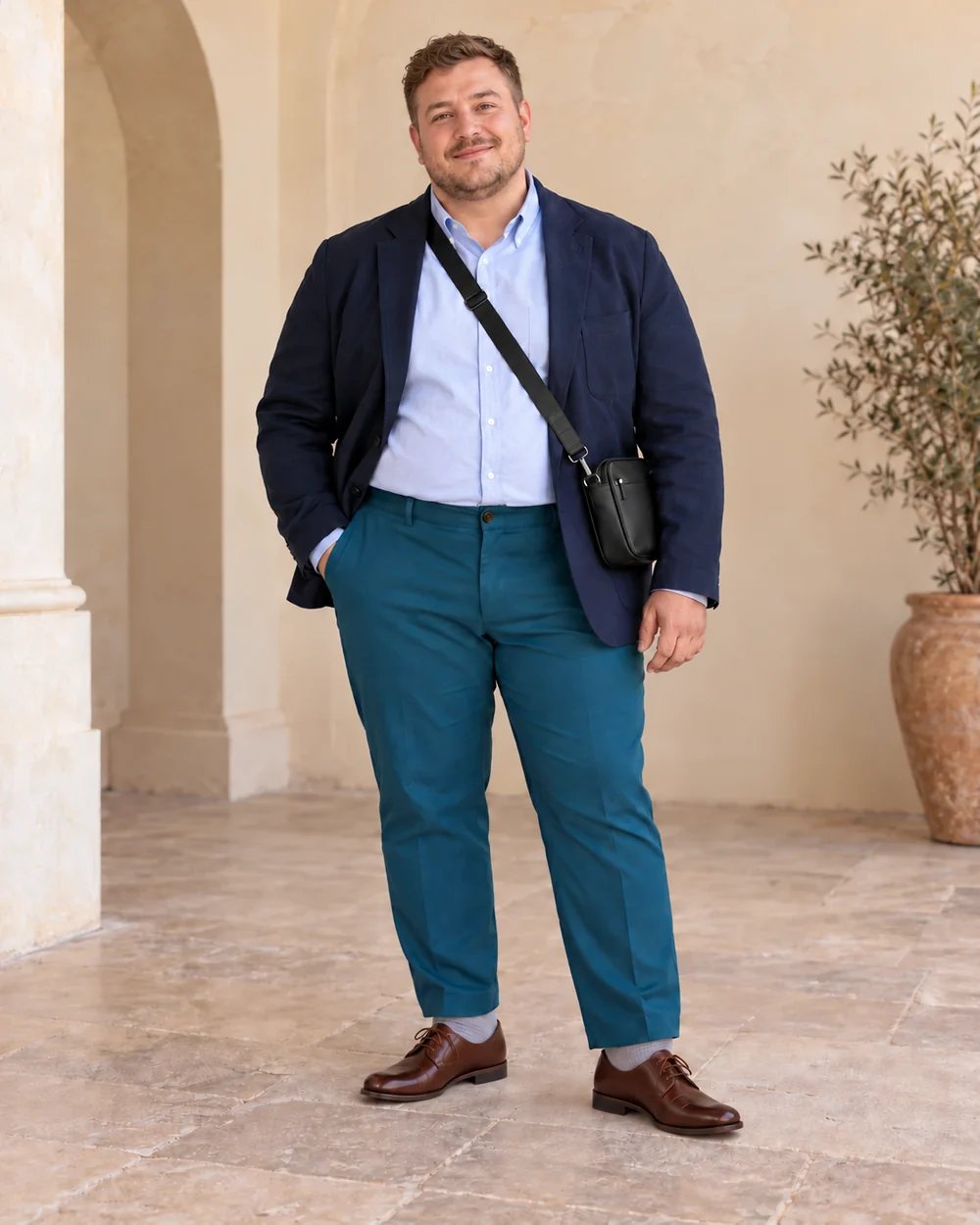 A fictional petite plus-size adult man with light-brown hair wearing teal tailored trousers, a pale-blue Oxford shirt, navy jacket and brown Derby shoes, with a dark crossbody bag.
