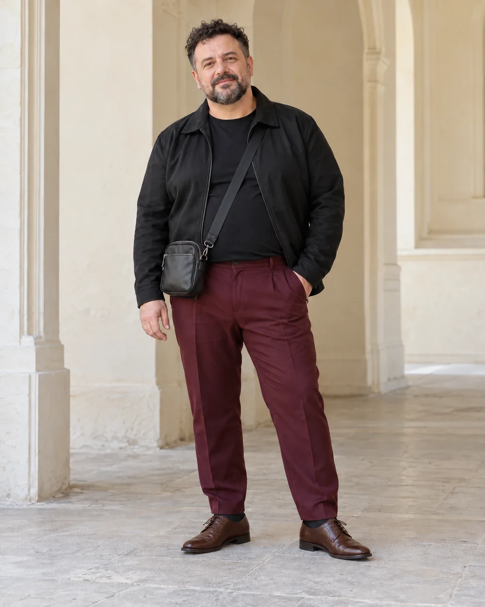 A full-length adult man with fair cool skin wearing Burgundy tailored trousers, black smooth tee, Black short jacket and Brown Derby shoes, with a small dark crossbody bag; both complete shoes are visible.